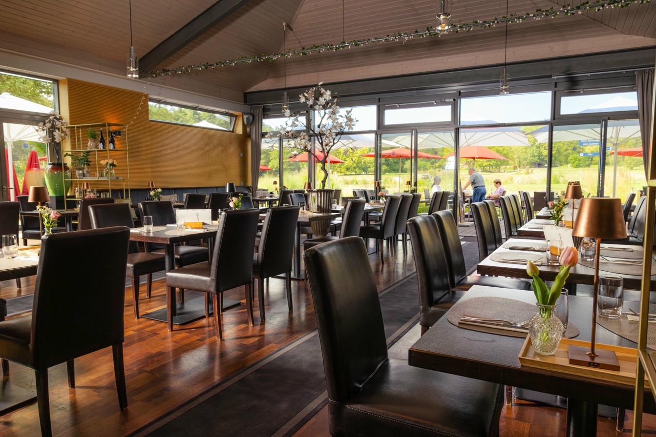 Restaurant-Innenraum mit hohen Decken, Parkett und Blick durch große Fenster auf den Golfplatz